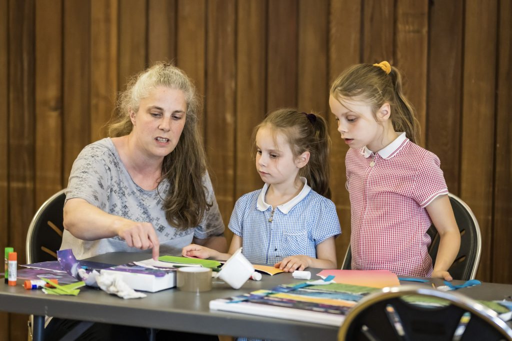 An adult and two children sit together at a table, creating an art work together. They are all looking thoughtfully at the materials on the table while they decide what to do next.