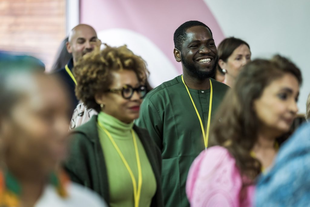 A man in a green tunic smiles broadly, standing in a crowd of diverse people wearing yellow lanyards.