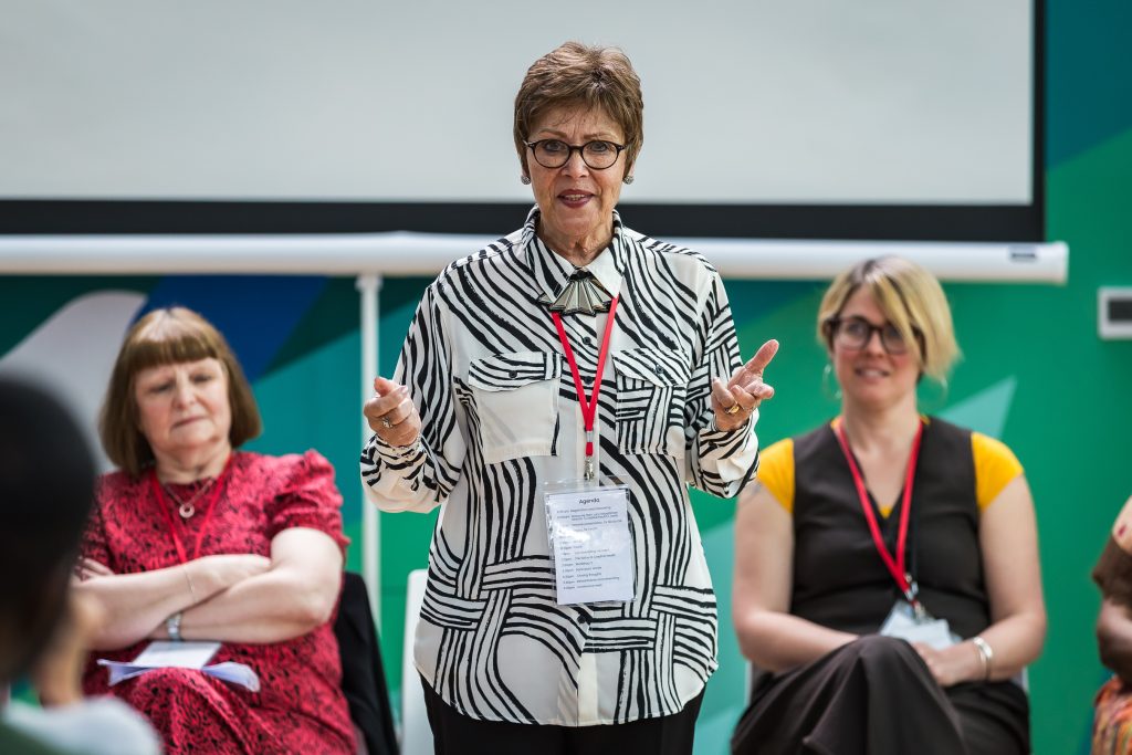 A woman in a black and white patterned shirt is gesturing with her hands while speaking at a conference.