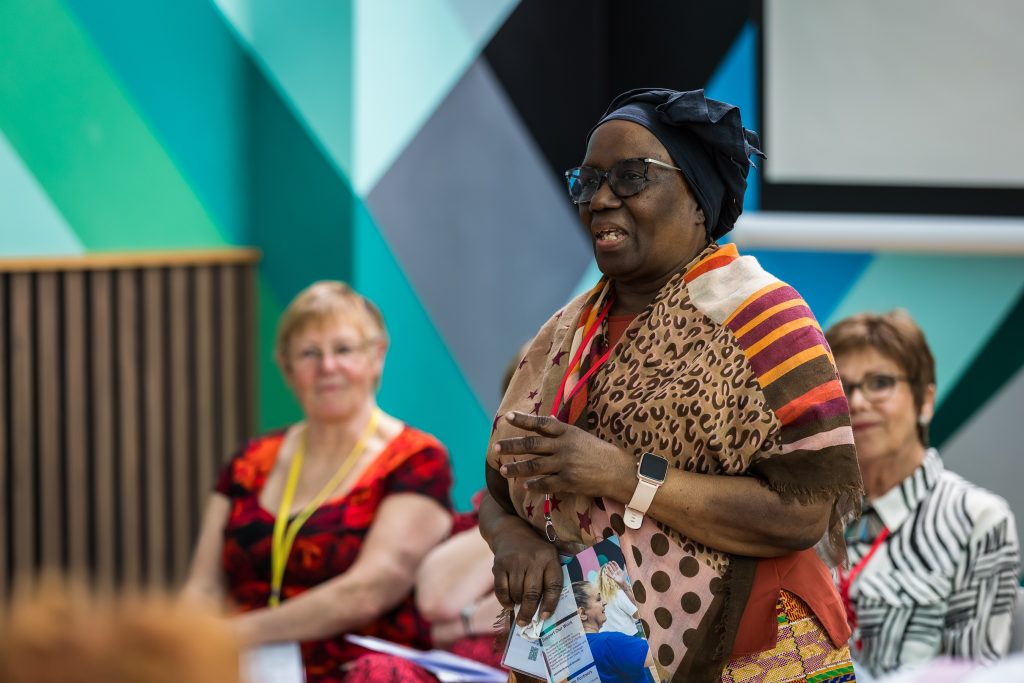 A woman wearing a black headscarf, colourful patterned scarf and dress gestures with her hands as she addresses an audience. Two other women are blurred in the background. The wall behind them features a geometric design of teal, green, and grey shapes.