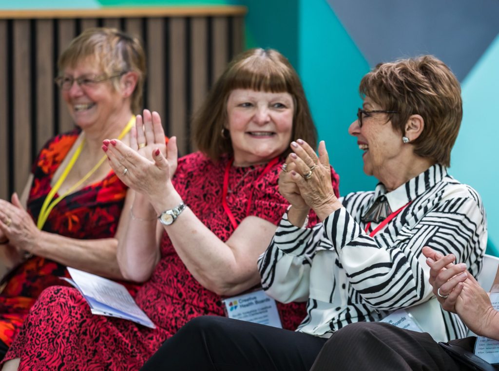 Three seated smiling people clap their hands. They are seated in front of a backdrop with geometric patterns.
