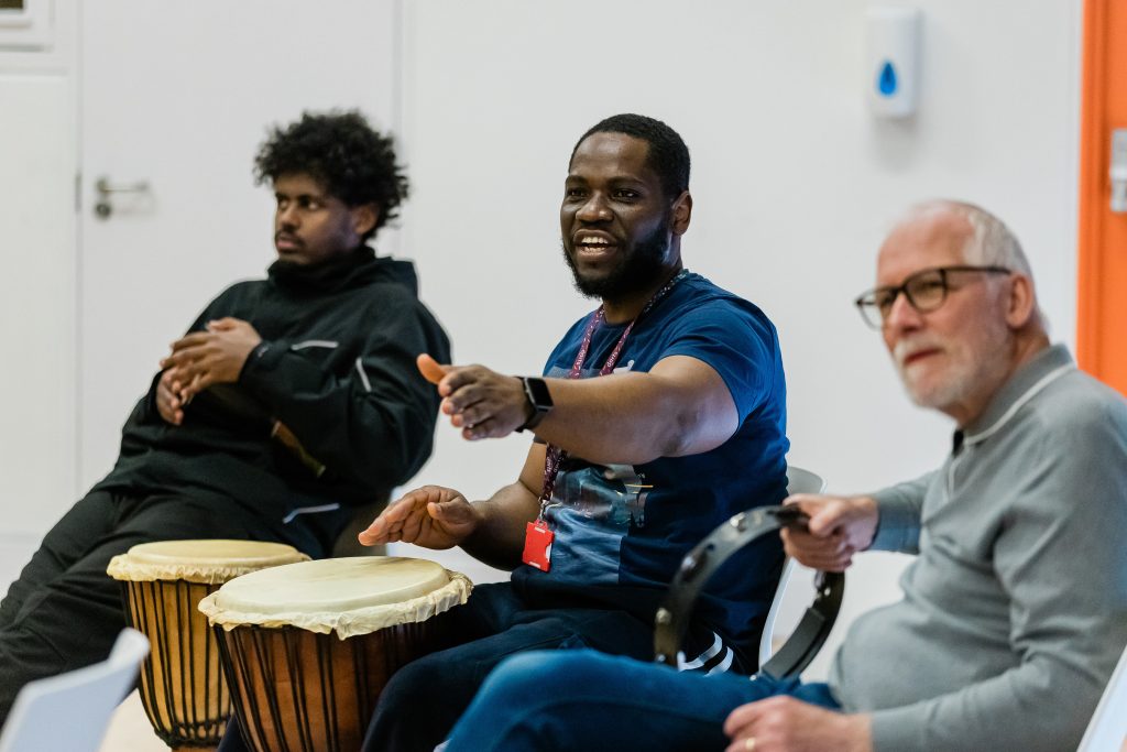 Three people of diverse backgrounds are seated in a room, engaged in a music activity. The central person is playing a djembe drum, gesturing with his left hand and smiling.