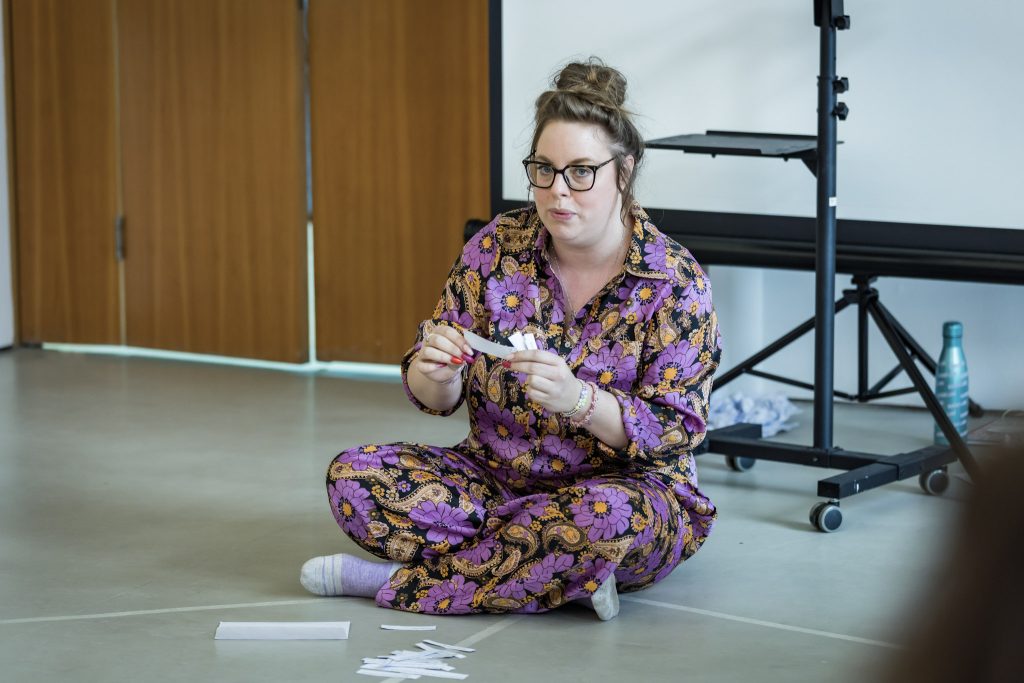 A person wearing a purple and black paisley patterned jumpsuit sits cross-legged on a light grey floor, holding several small white strips of paper.