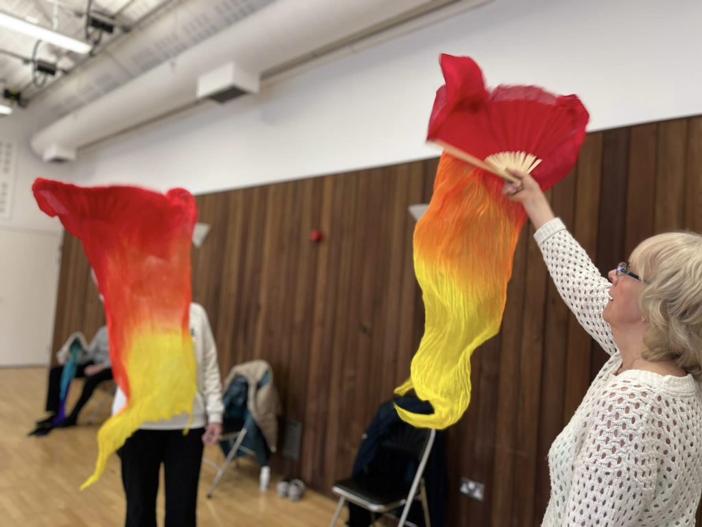 Two adults are waving flowing red and yellow scarves in a studio with wood-panelled walls.