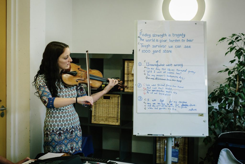 An adult with long dark curly hair wearing a patterned dress plays the violin next to a whiteboard of song lyrics and ideas.